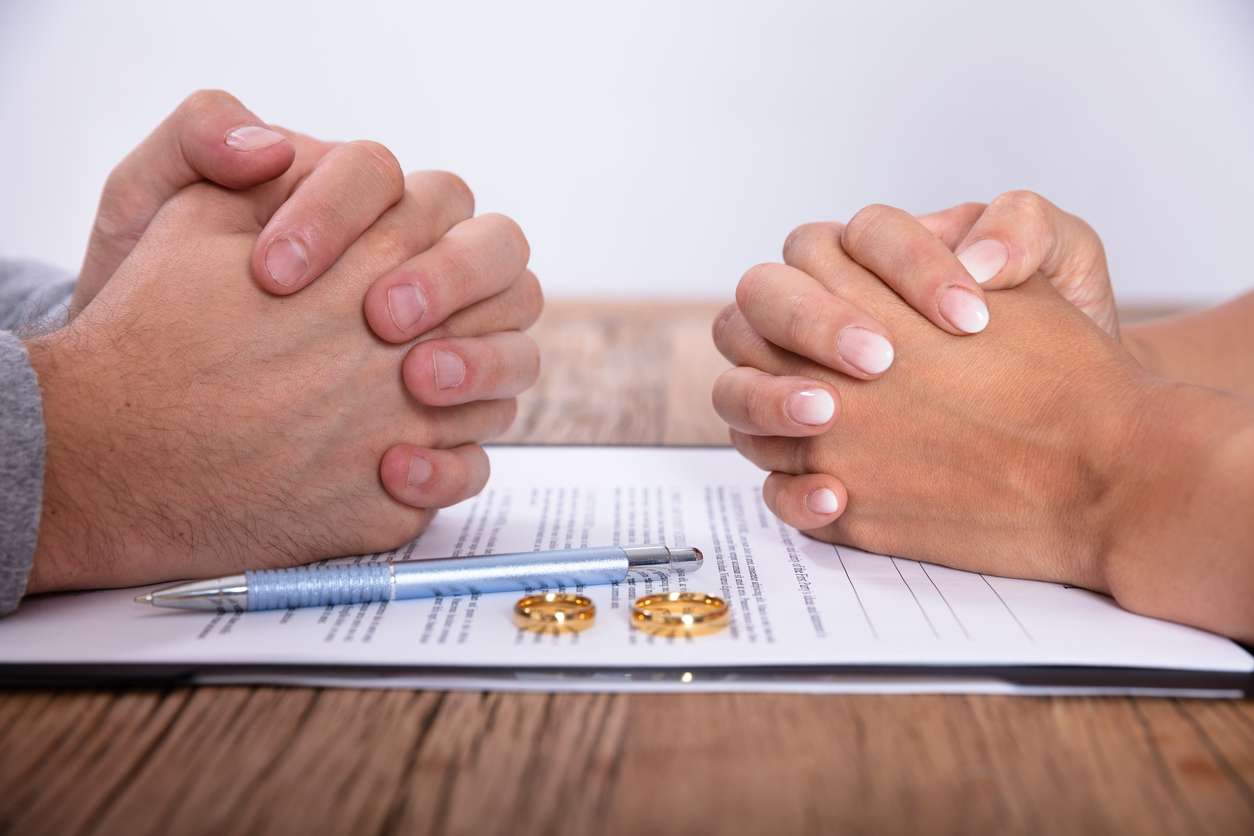 A close-up of two clasped hands over a document, with wedding rings nearby, suggesting a serious conversation or decision.